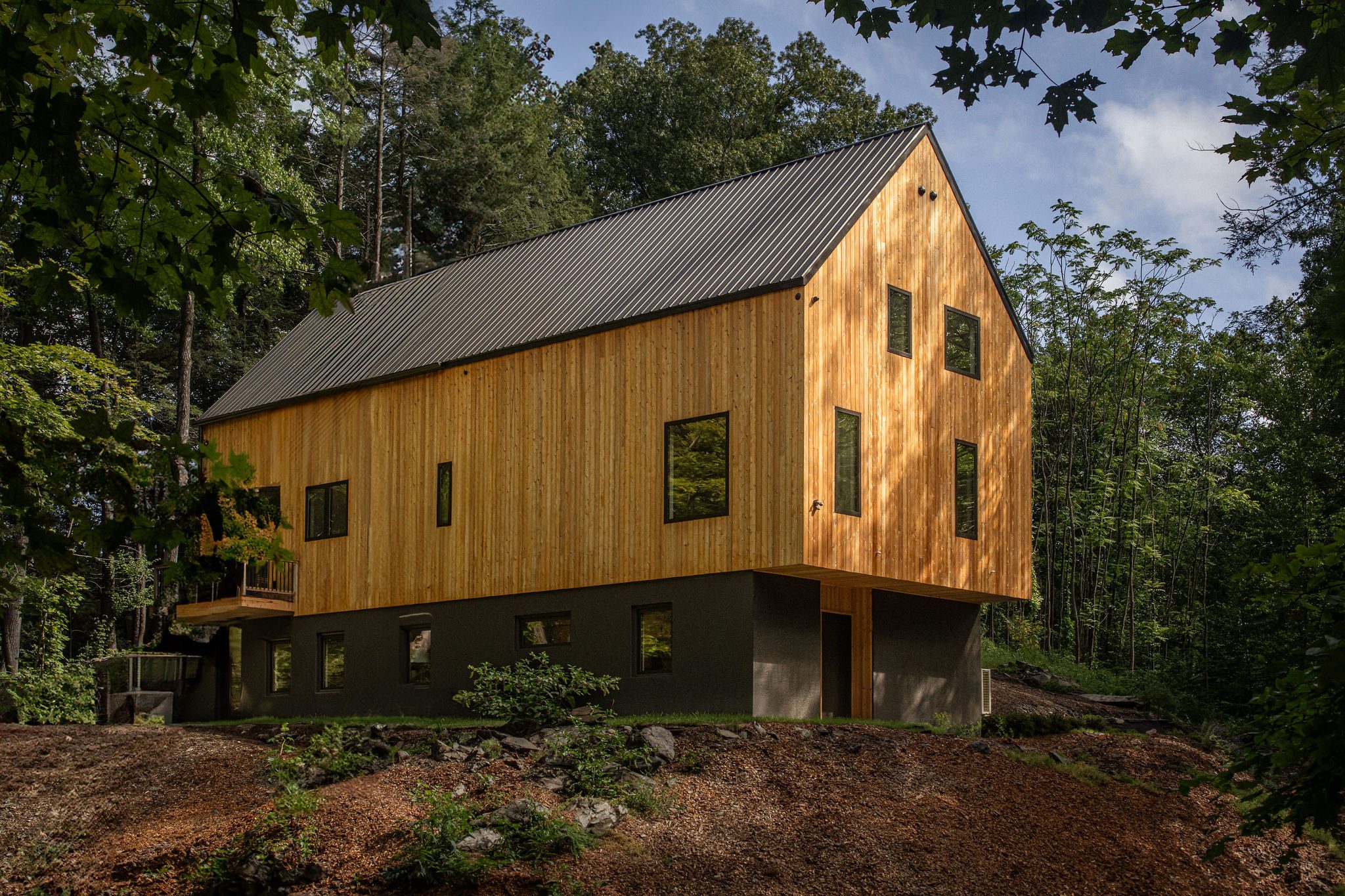 Cedar-clad house with cantilevered gable end over a dark base, set in woodland, Rhinebeck